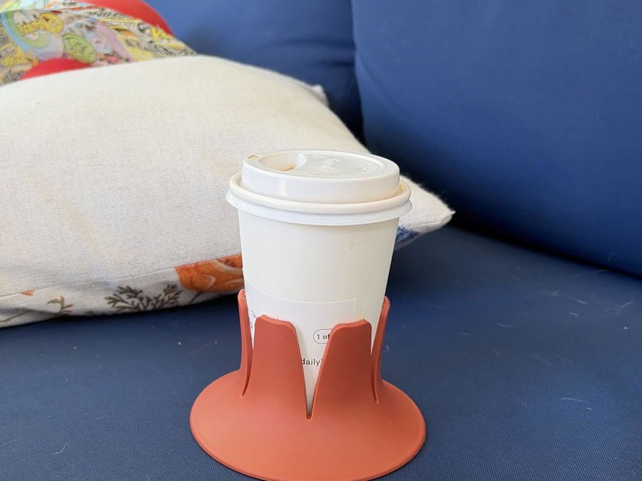A white paper coffee cup with a lid seated in a terracotta STEADI base on the navy cushion of an outdoor patio sofa, with a linen throw pillow and palm fronds behind in bright sun